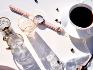 Gestão de Tempo: sem procastinar e com produtividade. Relógio perto de uma xícara de café, copos com água e grãos de café espalhados sobre a mesa.