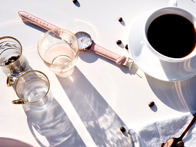 Gestão de Tempo: sem procastinar e com produtividade. Relógio perto de uma xícara de café, copos com água e grãos de café espalhados sobre a mesa.